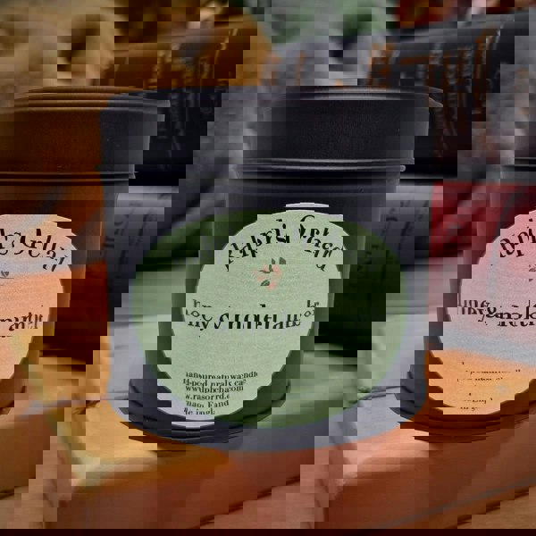 Candle tin labeled 'Ralph's Orchard honey & molten amber' on a wooden surface with books in the background.