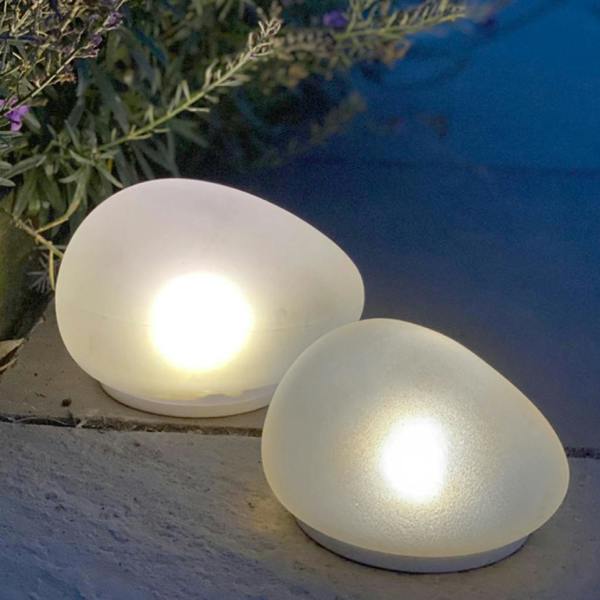 Two frosted glass solar powered pebble lights of different sizes glowing with warm white light on a stone garden patio at night.