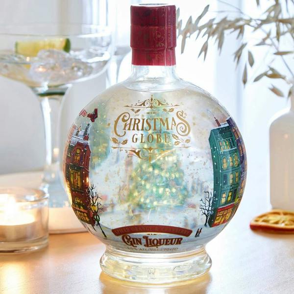 Illuminated Christmas globe gin liqueur bottle filled with gold leaf next to a lit candle and cocktail coupe.