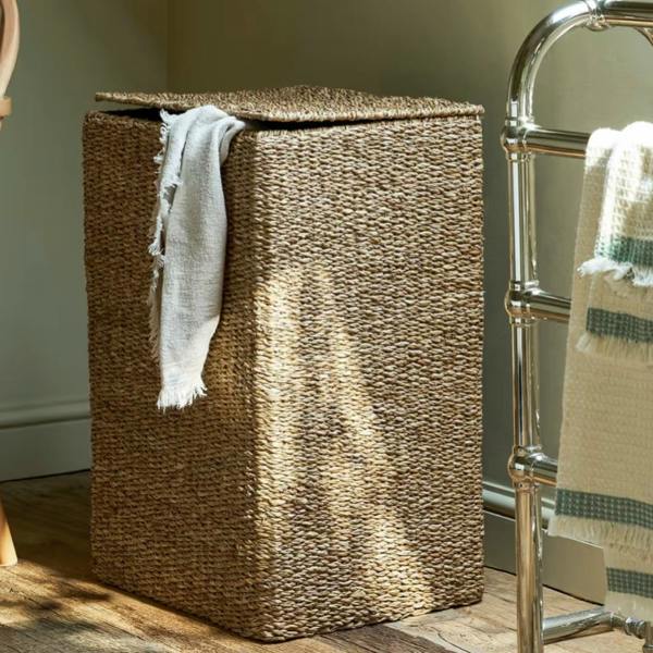 Rectangular handwoven seagrass laundry basket with a lid placed next to a heated towel rail in a sunlit bathroom.