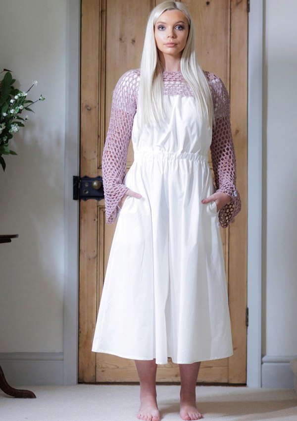 This is a young woman with long blonde hair wearing a Bardot style cream dress with short puff style sleeves. She is silhouetted against an internal room background and is facing the camera. She has her hands in her pockets and she has got bare feet. She is wearing a pink croched top over the dress.