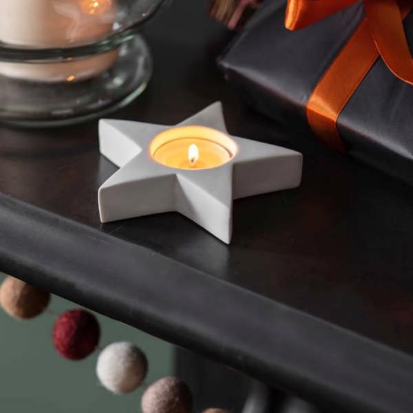 Two white star-shaped matte porcelain tealight holders on a dark mantelpiece with a lit candle.