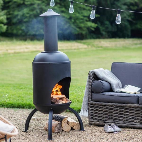 A Black Steel Chiminea with a lit wood fire, positioned on a gravel patio next to a modern grey garden sofa and outdoor string lights.
