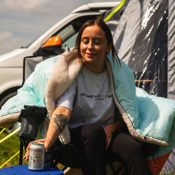 A young lady sat in a camping chair enjoying a drink whilst camping wrapped in an Insulated Outdoor Blanko by OLPRO in Tonal Treetops design. imageset:Tonal Tree Tops