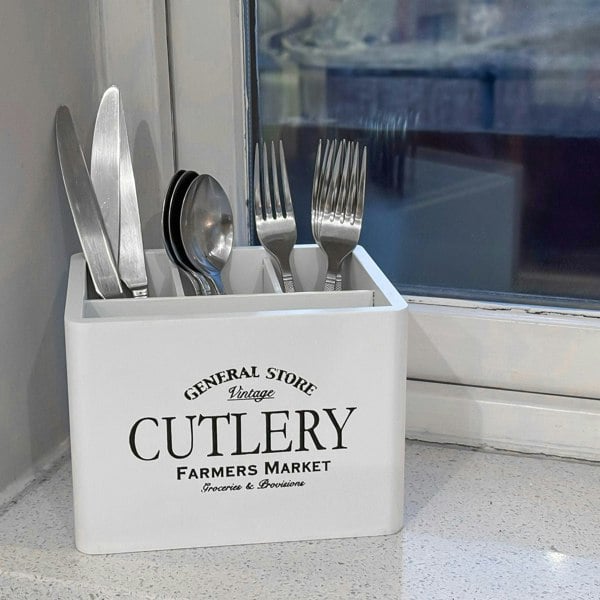 A white wooden cutlery caddy with vintage print, placed on a countertop, filled with various cutlery items such as knives, forks, and spoons.