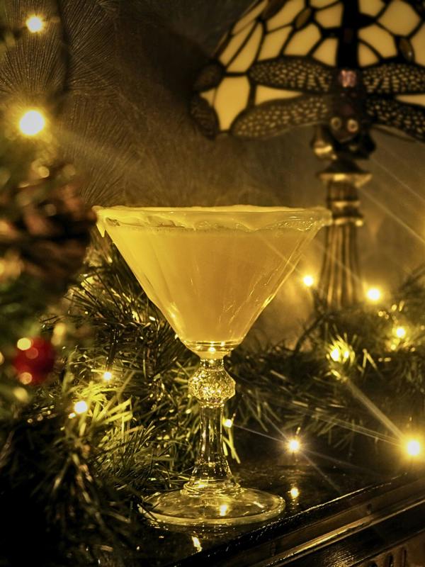 A clear martini-style coupe glass filled with a pale yellow drink, set on a dark tabletop with holiday greenery, warm string lights, and a stained-glass-style lamp in the background.