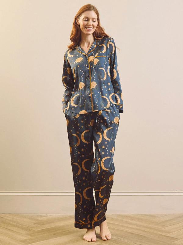A woman stands barefoot in a bedroom setting wearing a matching pyjama set with a dark blue base and a repeating celestial print of gold crescent moons,  stars and cats.