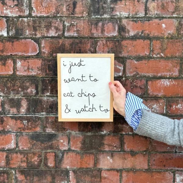 A hand wearing a grey wool jumper and blue striped shirt cuff holding a solid oak framed white cotton typography print displaying the lowercase handwritten-style quote "i just want to eat chips & watch tv" against a weathered red brick wall.