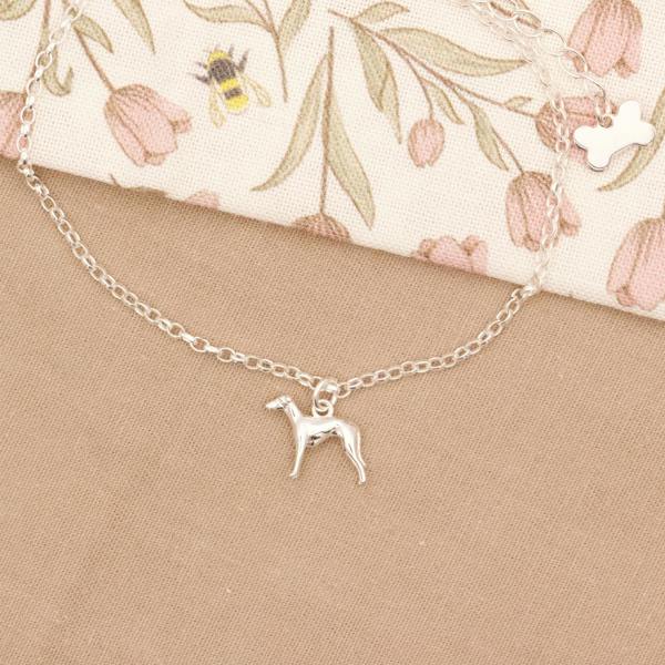 Silver whippet dog charm bracelet on a beige surface with a floral-patterned fabric background