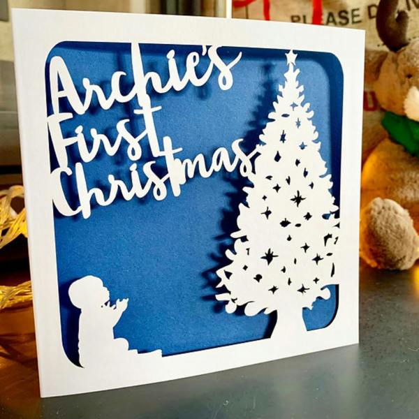 Personalised papercut first Christmas card featuring Archie's First Christmas, a baby silhouette, and Christmas tree against a blue paper insert