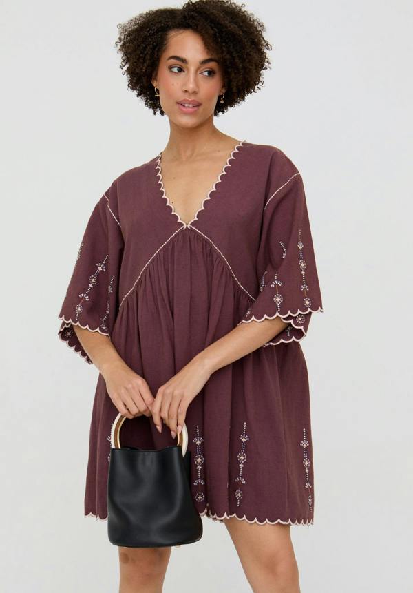 Woman wearing a burgundy dress with white embroidery, holding a black handbag on a light gray background