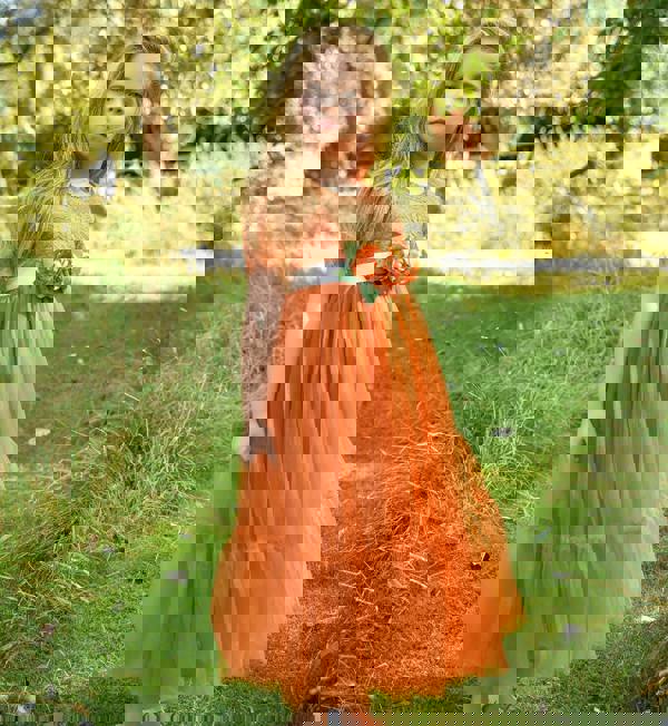 Young girl in an orange dress with a floral detail standing in a grassy outdoor setting.