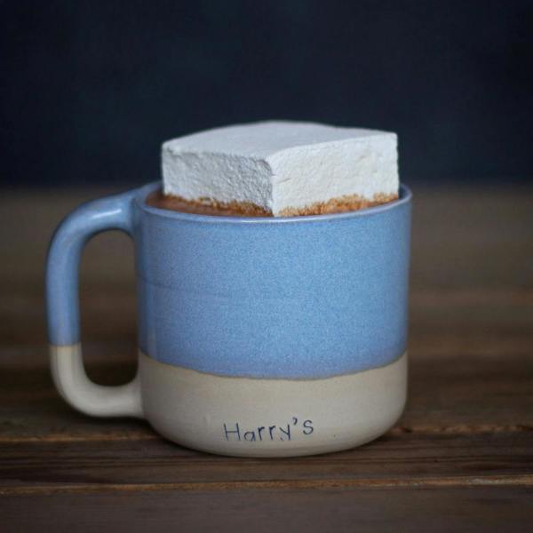 A blue and cream handmade ceramic mug filled with hot chocolate and topped with a large square marshmallow, resting on a wooden surface.