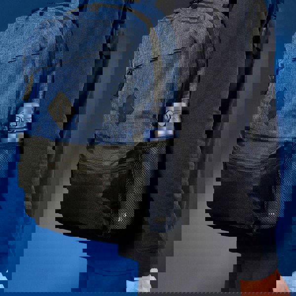 A blue backpack with a black pocket holds a blue water bottle in a mesh side pouch worn by a person dressed in dark clothing against a blue background.