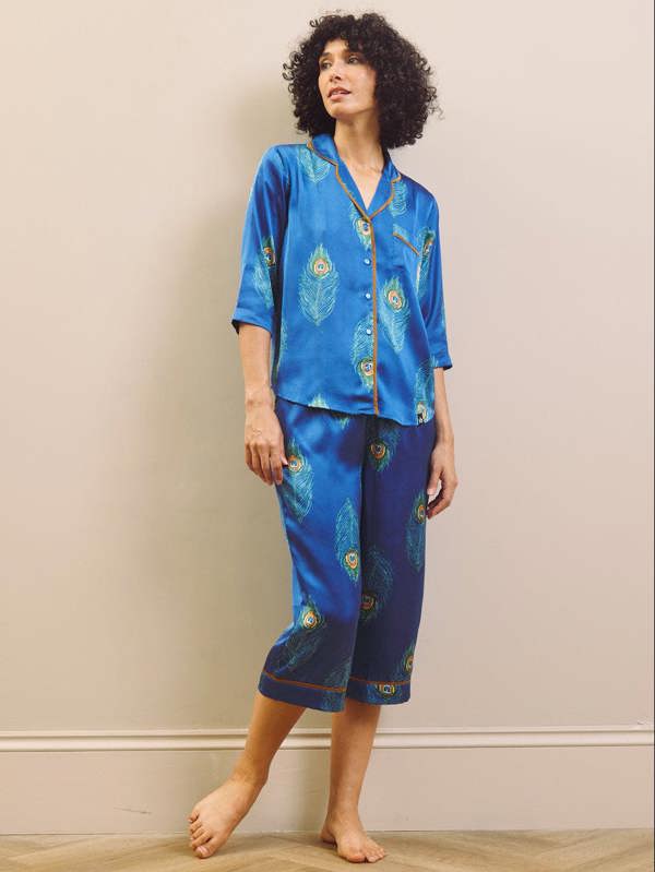 Woman wearing a blue pajama set with peacock patterns against a beige wall.