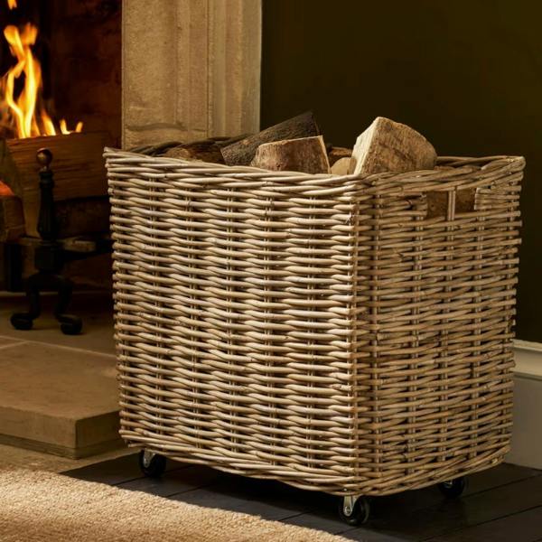 Large handwoven grey kubu rattan log basket on wheels filled with firewood sitting next to a roaring fireplace.