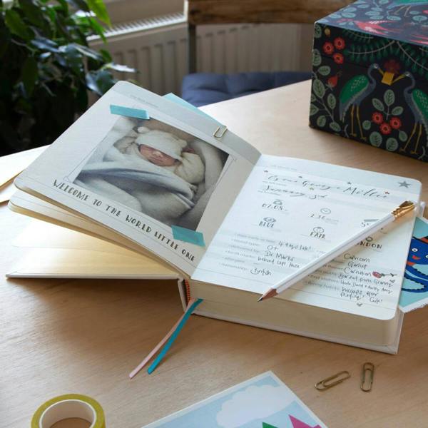 An open linen-bound baby journal sitting on a wooden desk, displaying a newborn photo on the left page and handwritten birth details on the right, accompanied by a pencil and paperclips.