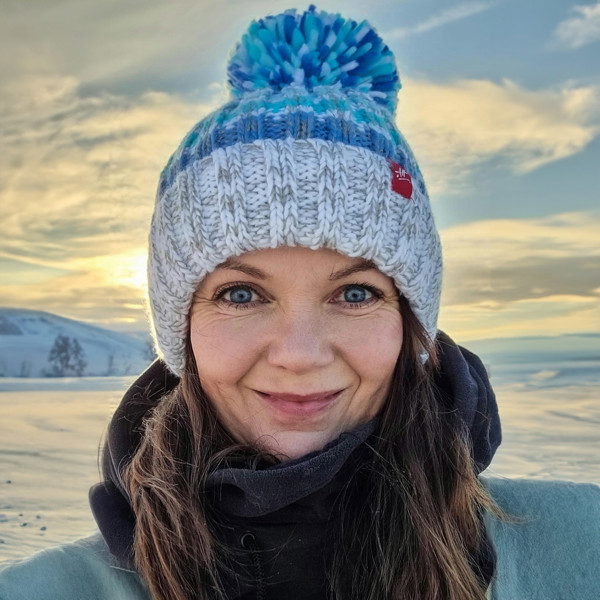  Woman against the backdrop of snow is wearing a Super Bobble hat with a base of white and pale grey and shades of sea blue stripes. The hat has a deep ribbed headband, contains reflective yarns andis lines in luxurious sherpa fleece.