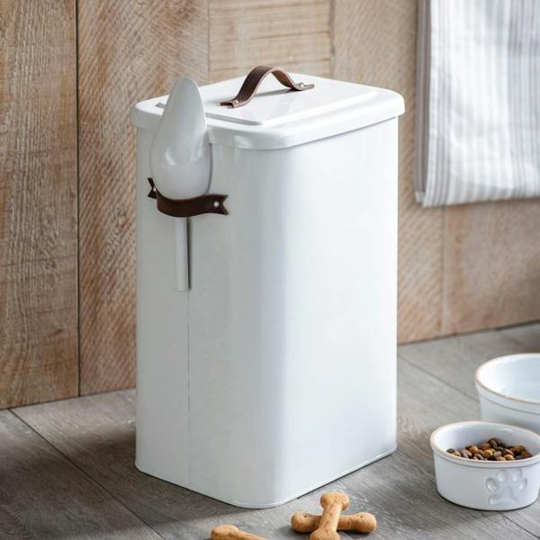 A tall, large white chalk-coloured powder-coated steel pet food bin featuring a brown leather lid handle and a side scoop, sitting on a wooden floor next to ceramic pet bowls and treats.
