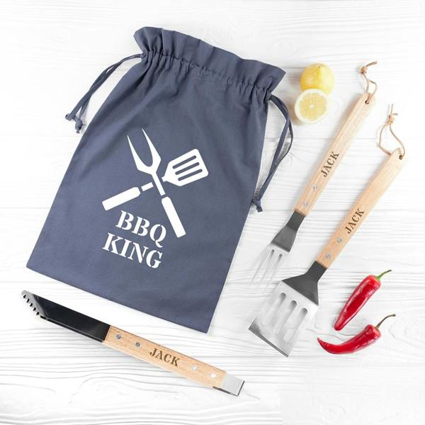 An overhead shot of three personalised BBQ tools with wooden handles engraved with the name Jack, resting next to a grey BBQ King drawstring bag on a white wood backdrop.