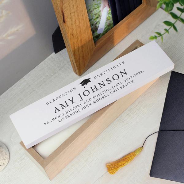 A personalised graduation wooden certificate box with a white lid showing a graduate's name, BA history and politics honours degree, and university text, styled next to a wooden photo frame.