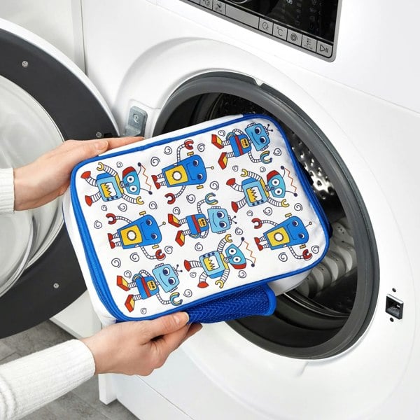 Hands are placing a colorful pouch decorated with blue robots into an open front-loading washing machine in a bright laundry room.