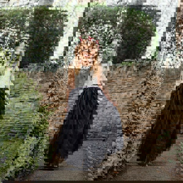 Young girl in a white top and black skirt with a flower crown standing outdoors.