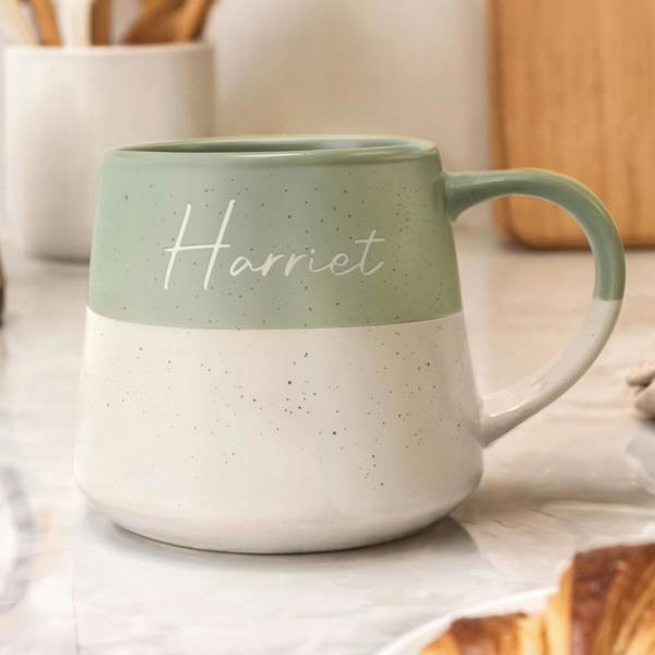 Personalised green and white speckled ceramic mug featuring the name "Harriet" in white script font on a marble kitchen worktop.