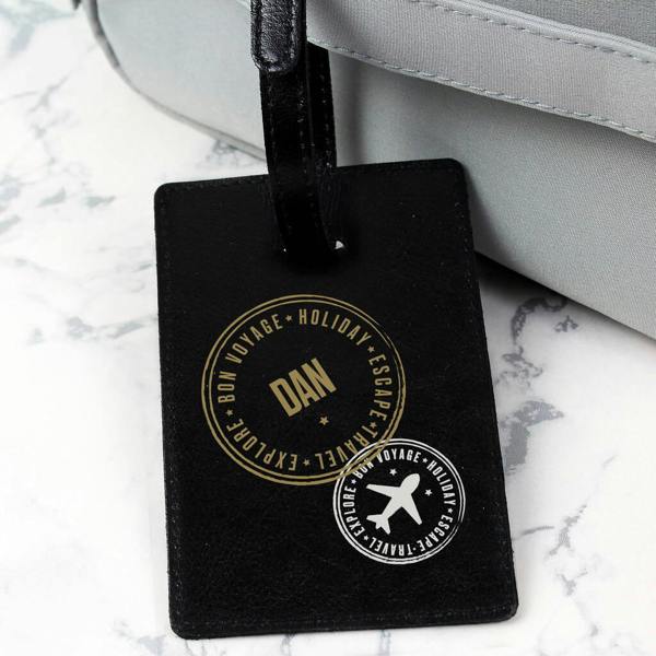 A black leather luggage tag featuring gold and white stamped designs, including an airplane icon and personalised name, attached to a grey travel bag.