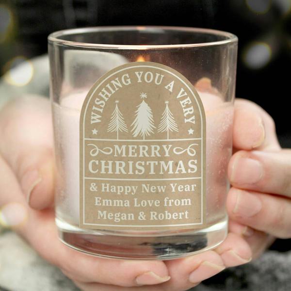 A person holding a glass Christmas jar candle featuring a festive tree arch label and a personalised holiday message.