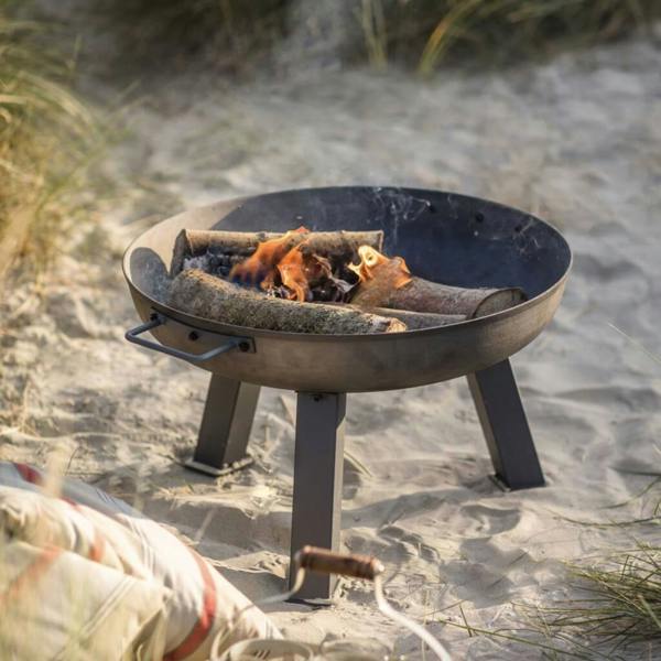 Small 55cm cast iron garden fire bowl with a lit fire, styled on a sandy beach with outdoor cushions in the foreground.