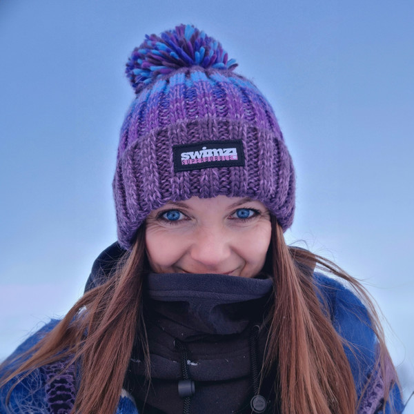  Woman standing in a snowy landscape in Norway is wearing a chunky knit Super Bobble hat in berry shades of purple and blue. The hat has a big pom-pom and a deep ribbed headband. It contains reflective yarns and is lined in luxury sherpa fleece. 