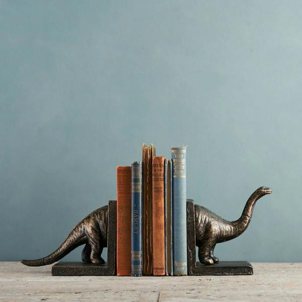 A pair of heavy-duty, bronze-coloured cast iron Brontosaurus bookends supporting a small row of vintage books on a wooden surface against a muted blue-grey wall.