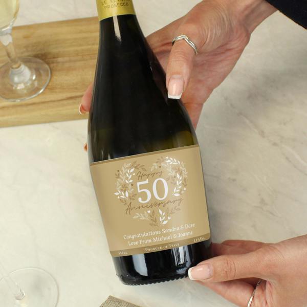 A close-up shot of a person holding a bottle of personalised 50th Golden Wedding Anniversary Prosecco from Abode Home & Gifts, featuring a custom gold label with text reading Congratulations Sandra & Dave Love From Michael & Joanne.