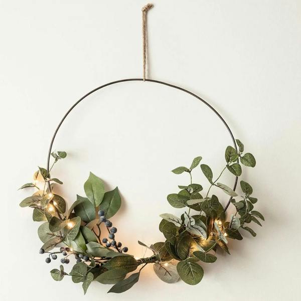 A minimalist 35cm Christmas half wreath made of a black metal hoop with artificial green eucalyptus, dark blue berries, and glowing warm white micro lights on a white wall.