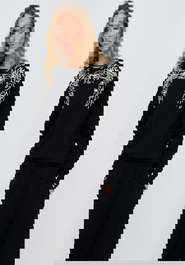 Woman wearing a black outfit with white floral embroidery on a plain background