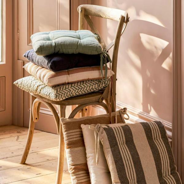 A stack of neutral and patterned linen seat pads, including Carbon and Rosemary, on a wooden dining chair.