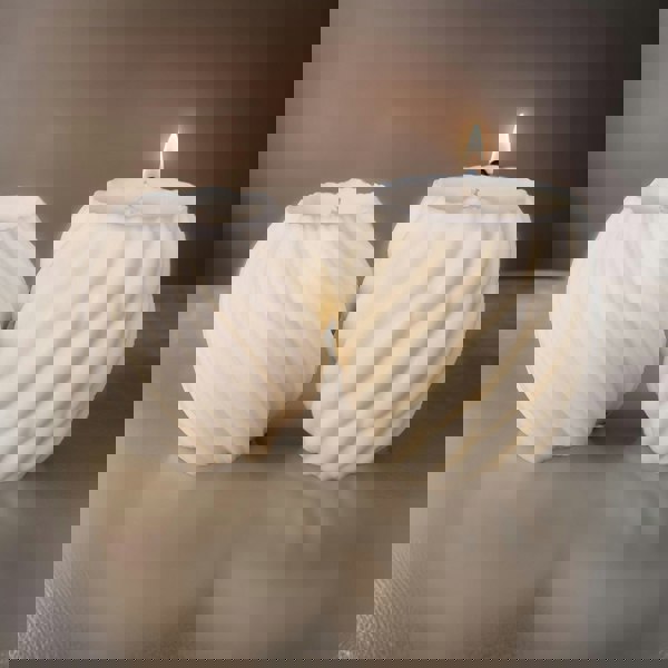 A white scented pillar candle with a twisted design, burning on a metal holder with a dark background.