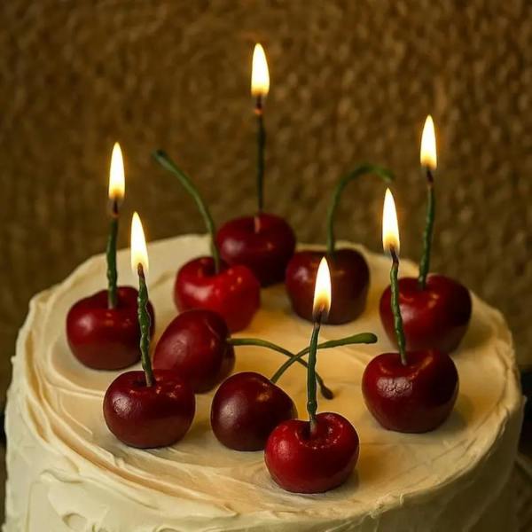 A close-up of eight lit, realistic red cherry-shaped candles with green stems arranged as toppers on a frosted white birthday cake.