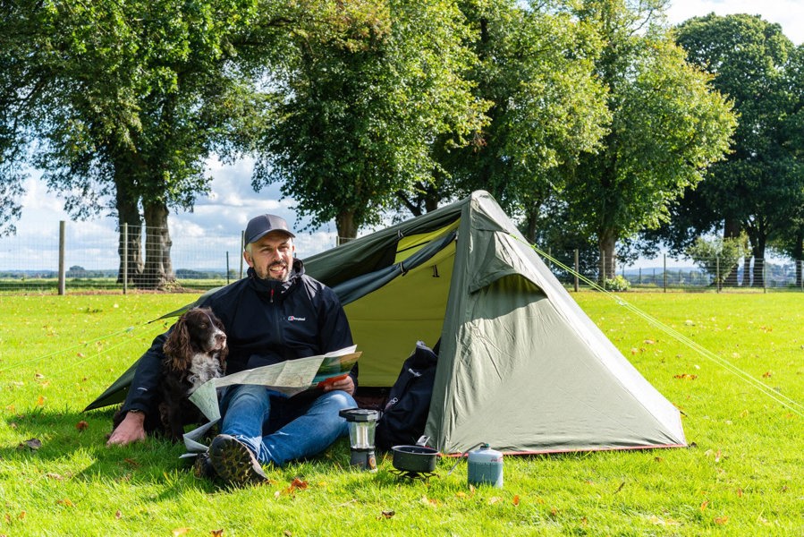 A walker and his dog look at a map in the OLPRO Hawford Lightweight 2 Person Tent pitched in a field with a tree line in the background.