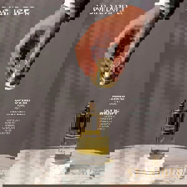 Hand holding a gold cap above a perfume bottle labeled 'Sapil Dapper' on a marble surface with a gray background.