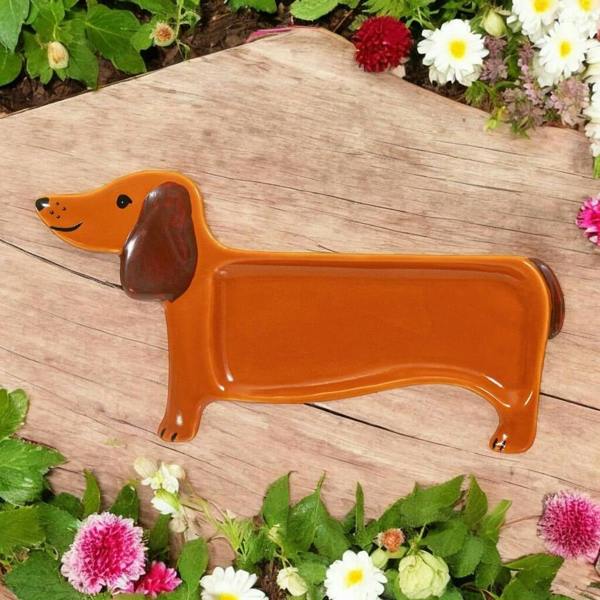 A hand-painted ceramic sausage dog tea bag rest with brown ears, displayed on a wooden board amongst garden flowers.