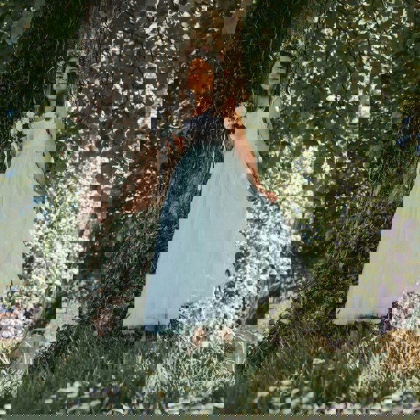 Young girl in a white and light blue dress standing under a tree with greenery around.