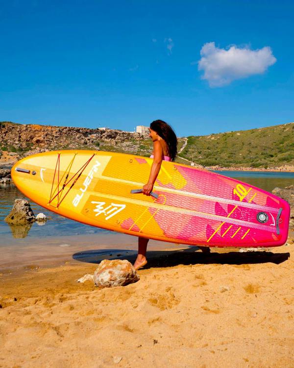 Woman walking on the beach holding a  Pink Coral 10' Lite Bluefin sup paddle board 