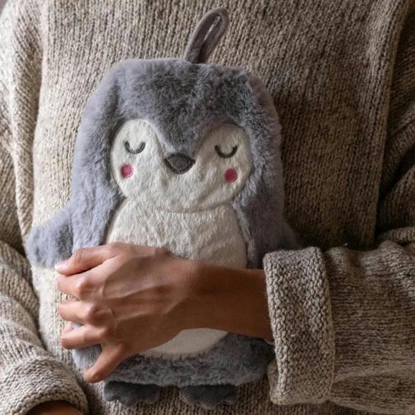 A person in a beige knit jumper holding a soft grey and cream plush penguin hot water bottle with pink rosy cheeks.