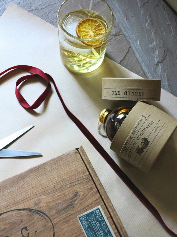A styled flat-lay of a bottle of Old Ginger by Prohibition Cocktails with kraft paper labels, a glass of the golden gin with a dried orange slice, scissors, a wooden box, and a red ribbon on a neutral background.