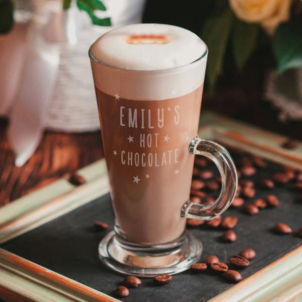 A tall clear glass filled with hot chocolate and whipped cream, engraved with "Emily's Hot Chocolate" surrounded by stars, on a rustic tray.
