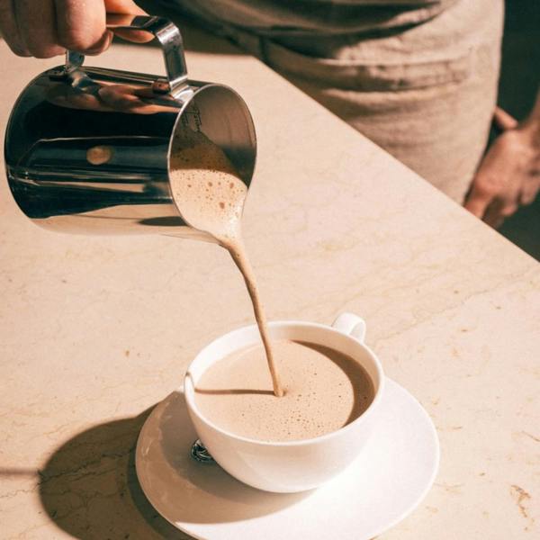 A side view of frothy, thick hot chocolate being poured from a stainless steel milk frothing pitcher into a white ceramic teacup and saucer.