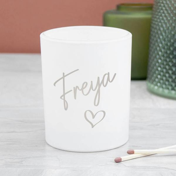 A white frosted glass candle holder printed with the name 'Freya' and a small heart icon sitting on a grey surface next to two matches.