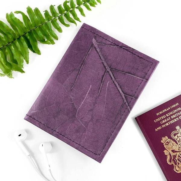 A bold purple Leaf Leather Passport Cover showcasing organic vein patterns, laid on a clean white surface next to a green fern leaf and a pair of white earphones.